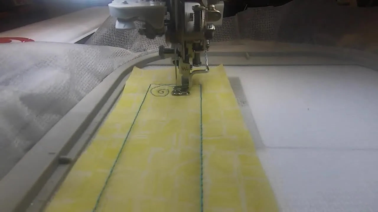 A close-up of the machine stitching the turquoise swirl decoration onto the yellow Halloween Town sashing strip in the hoop.