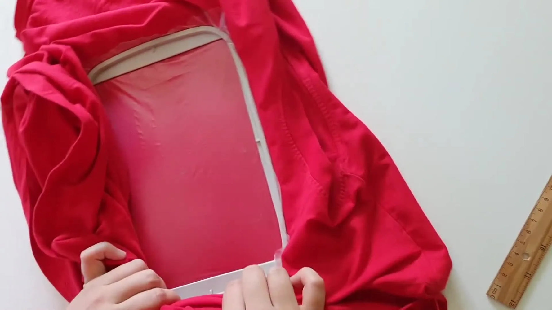 Hands gently tension a hooped red t-shirt until it feels drum-tight, the key moment that prevents knit distortion and puckering.