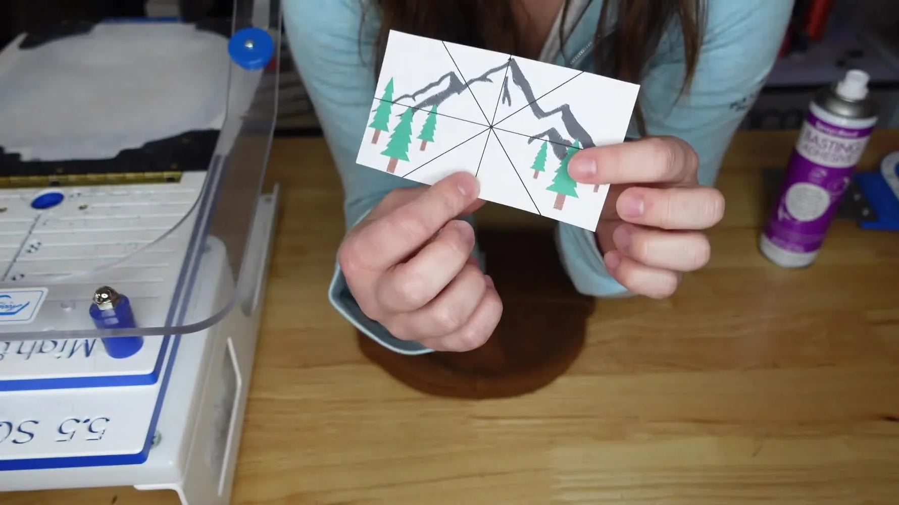 Hands holding a printed embroidery design showing a mountain and trees with a marked center point.