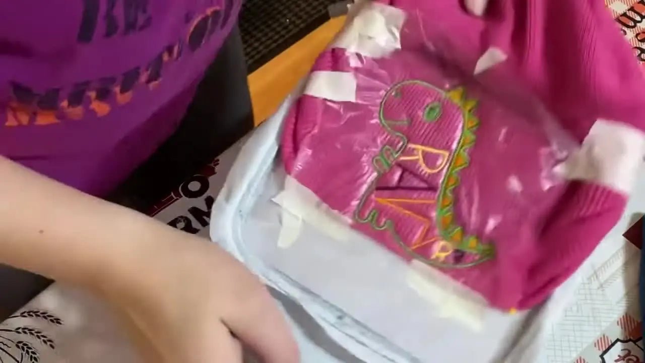 A finished dinosaur-and-text embroidery on a pink rimless beanie sits in the hoop under a clear water-soluble topper, showing crisp stitches on stretchy knit.