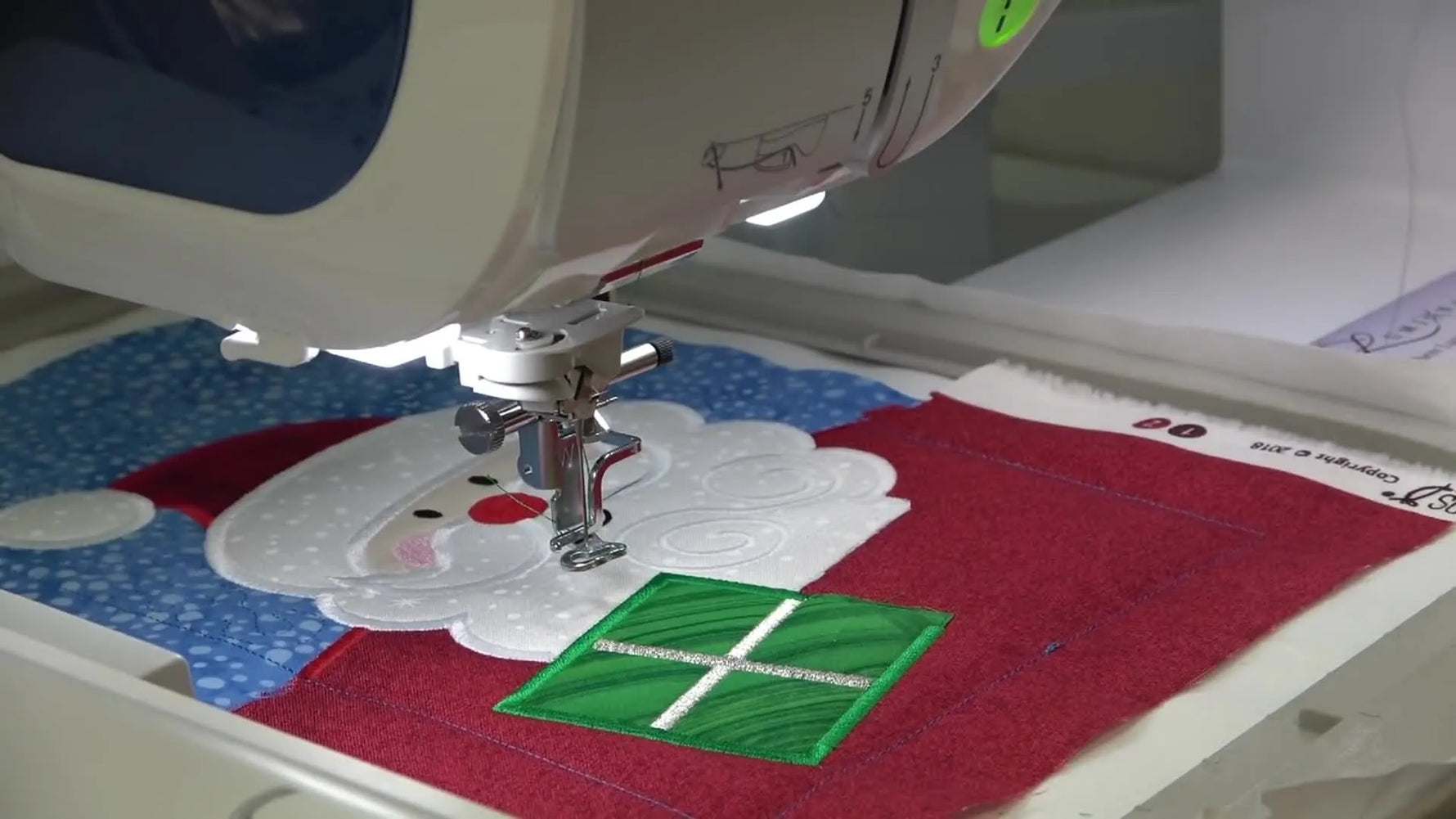 The finished Sweet Pea Santa #4 appliqué is held up for a final reveal, showing clean satin edges and metallic bow details.