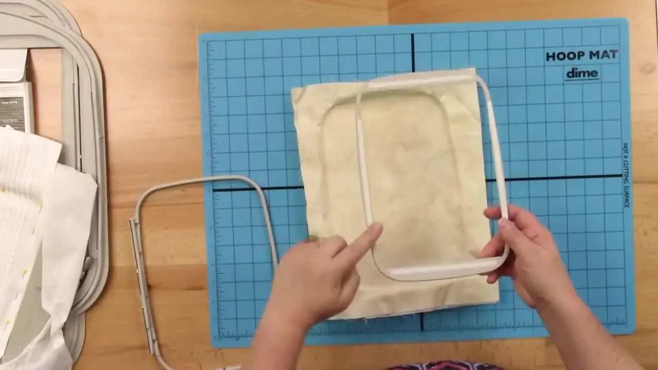 Hands peel the backing off double-sided Wonder Tape applied to the underside of an inner embroidery hoop ring to stabilize fabric during hooping.