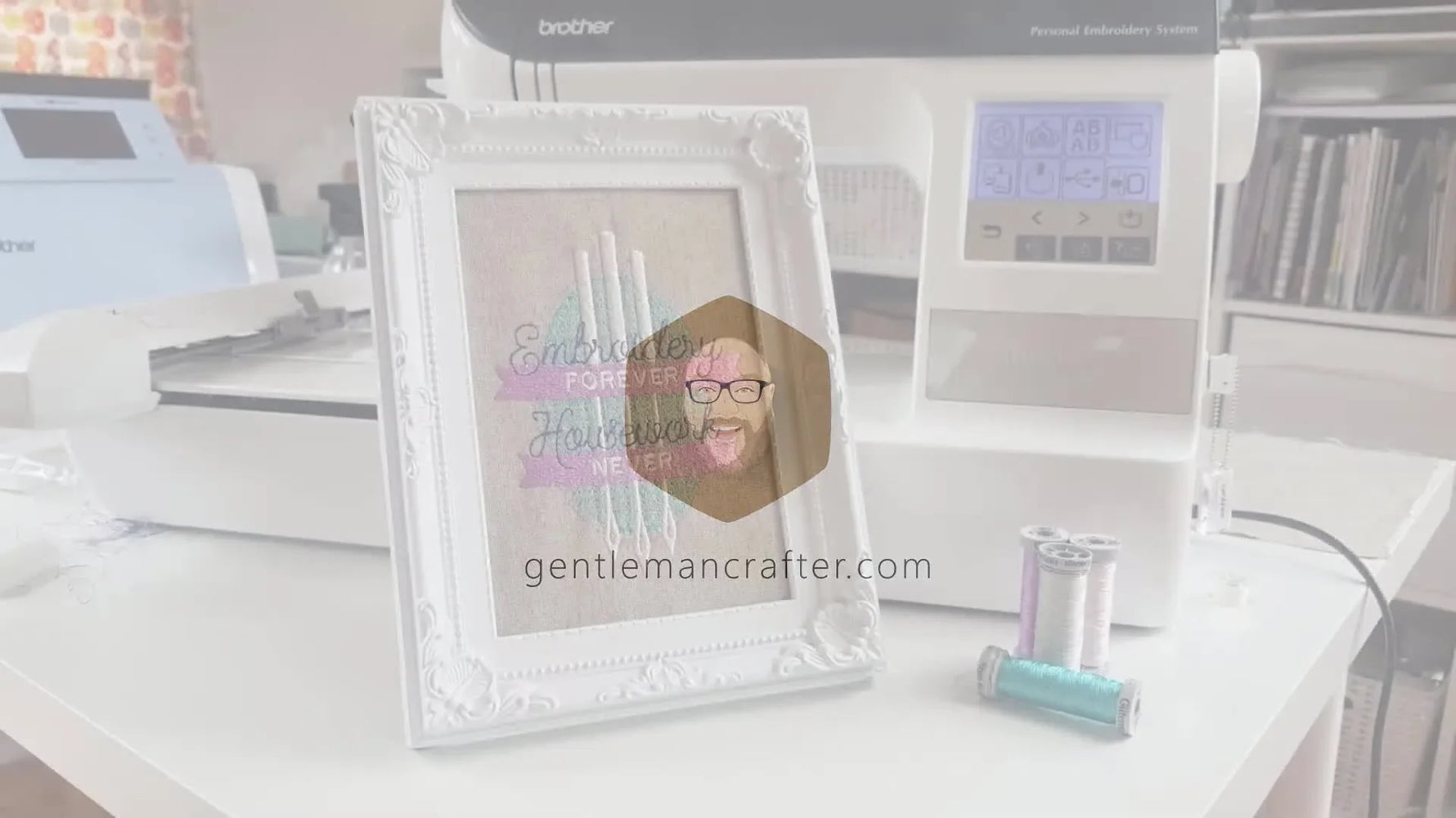 Finished embroidered design in a white ornate frame next to an embroidery machine and spools of thread.