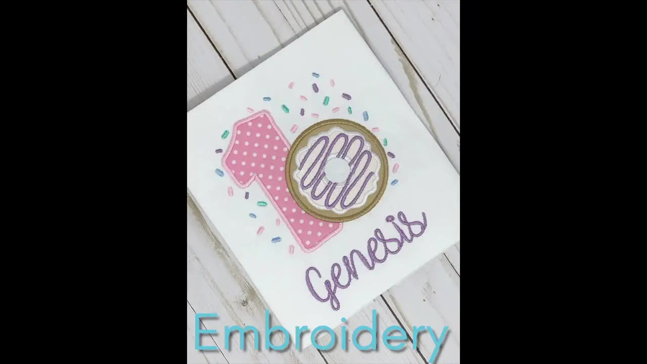 A white baby shirt with an embroidered '1' and donut for a first birthday, with 'Genesis' embroidered below.