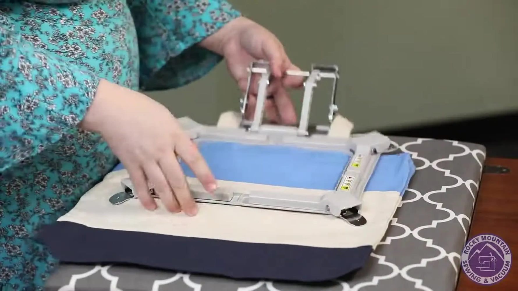 The presenter holds up a fully hooped canvas tote bag secured in the Brother 5x7 Versatile Magnetic Frame to show how firmly it clamps.