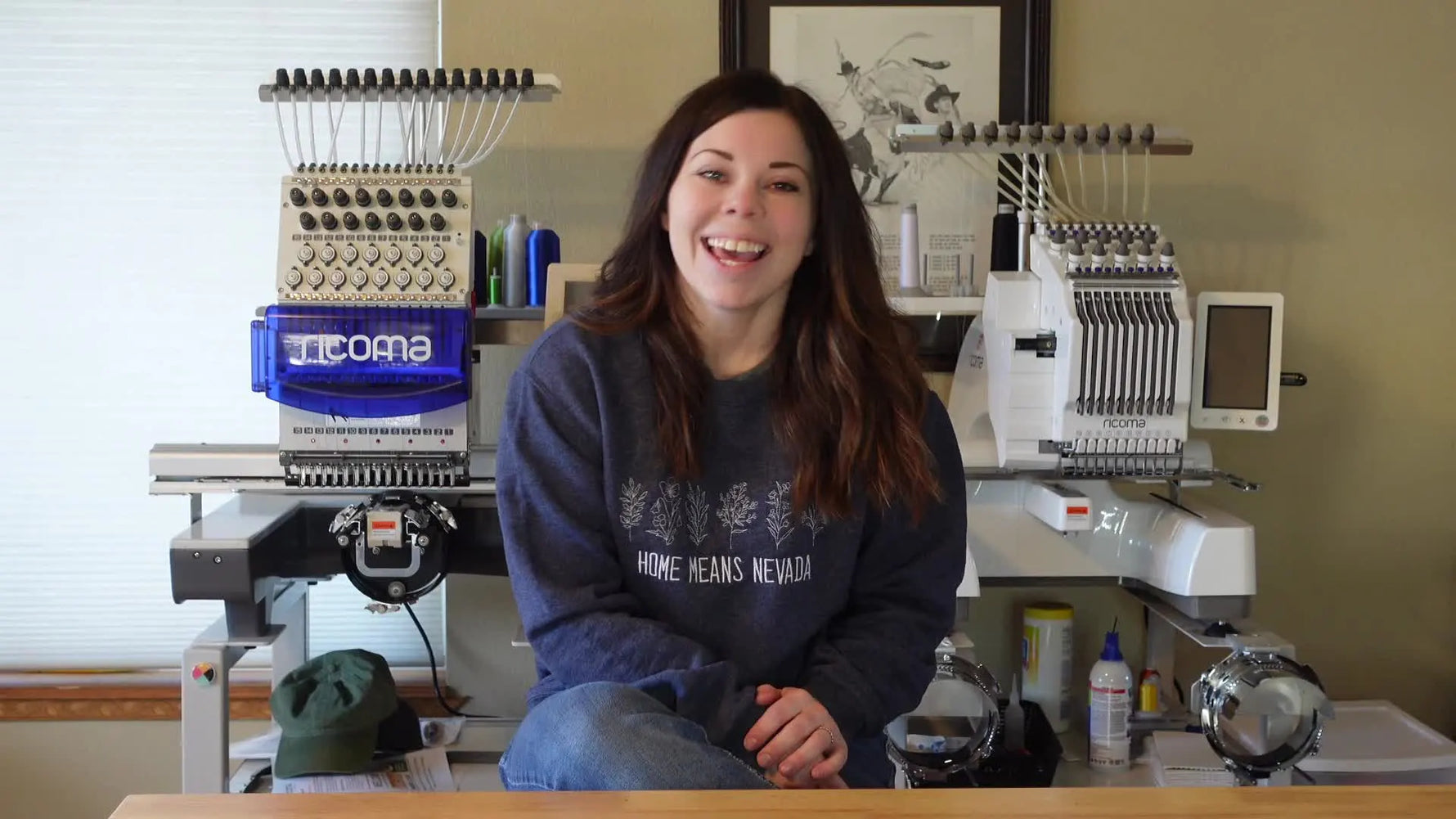 A wide studio shot showing Kayla seated between multi-needle embroidery machines, setting the context for beginner tips and workflow upgrades.