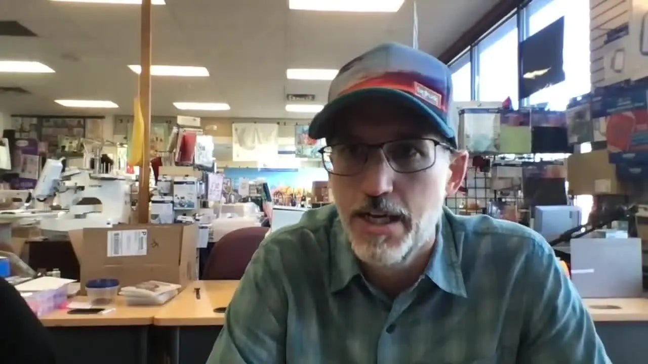 A sewing shop owner speaks directly to camera during a live stream, sharing shipping updates and timelines for Brother upgrades and 7x12 magnetic hoops.