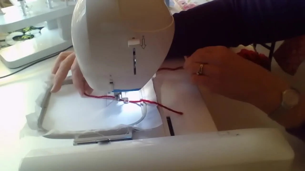 Close-up of a home embroidery machine zigzag-stitching over red yarn while a hand guides the yarn along the placement line.