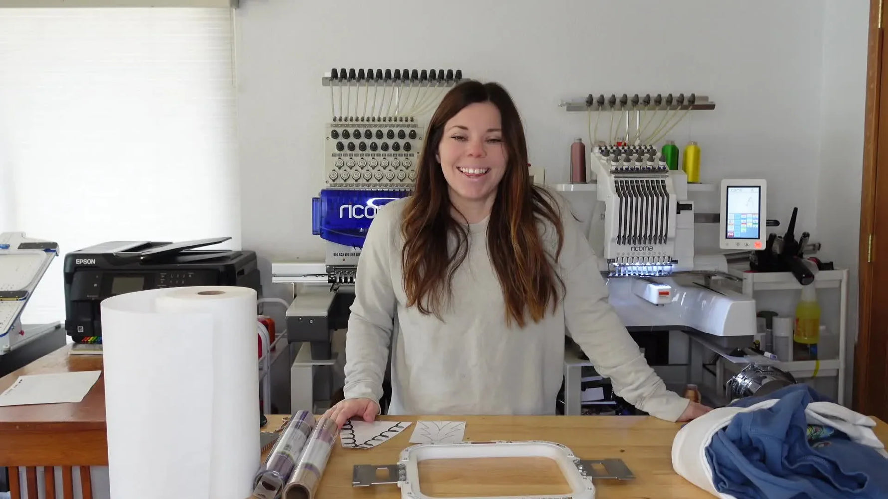 Woman presenting various embroidery materials on a wooden table, with embroidery machines in the background.