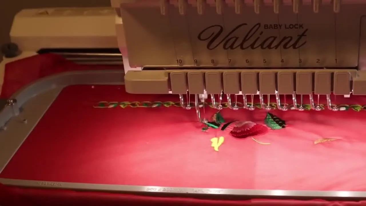 A close-up of a Baby Lock Valiant 10-needle embroidery machine stitching green leaves onto red silk georgette held in a large hoop.