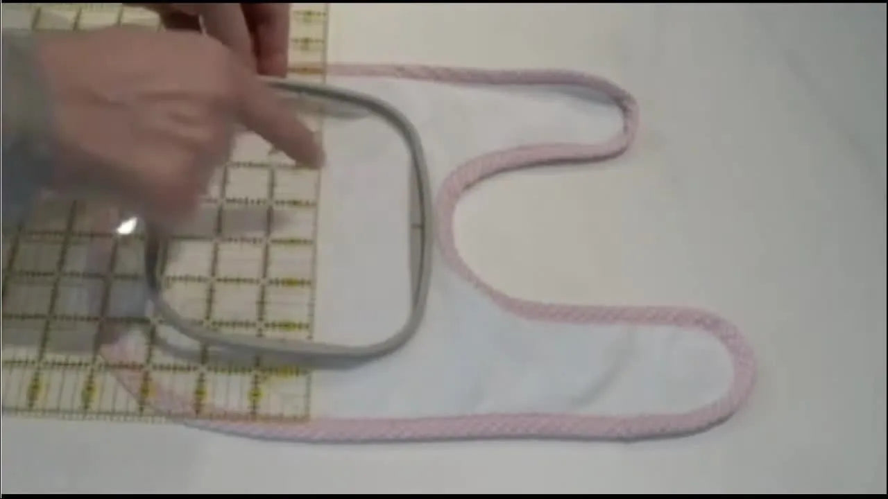An inner embroidery hoop is aligned to a clear quilting ruler grid on a baby bib for precise, no-shift placement before hooping.