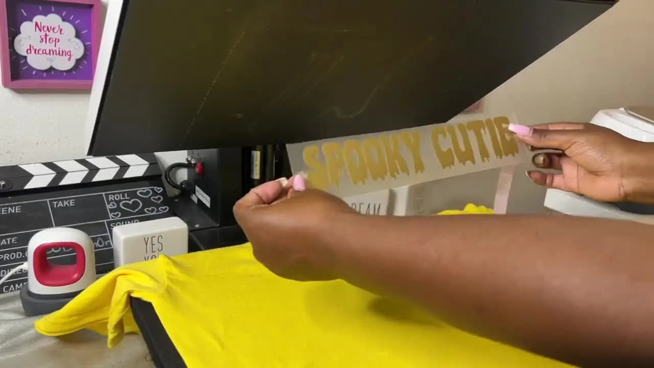 The finished yellow T‑shirt is held up to show the embroidered ghost-and-pumpkin design layered with raised metallic gold puff vinyl text.