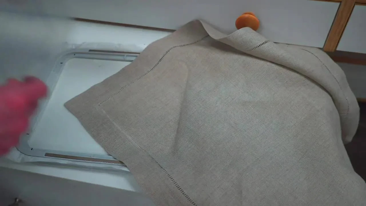 A clear plastic grid template sits over a hooped linen napkin so the hidden RE18 hoop reference marks can be extended onto the fabric for accurate AcuSetter positioning.