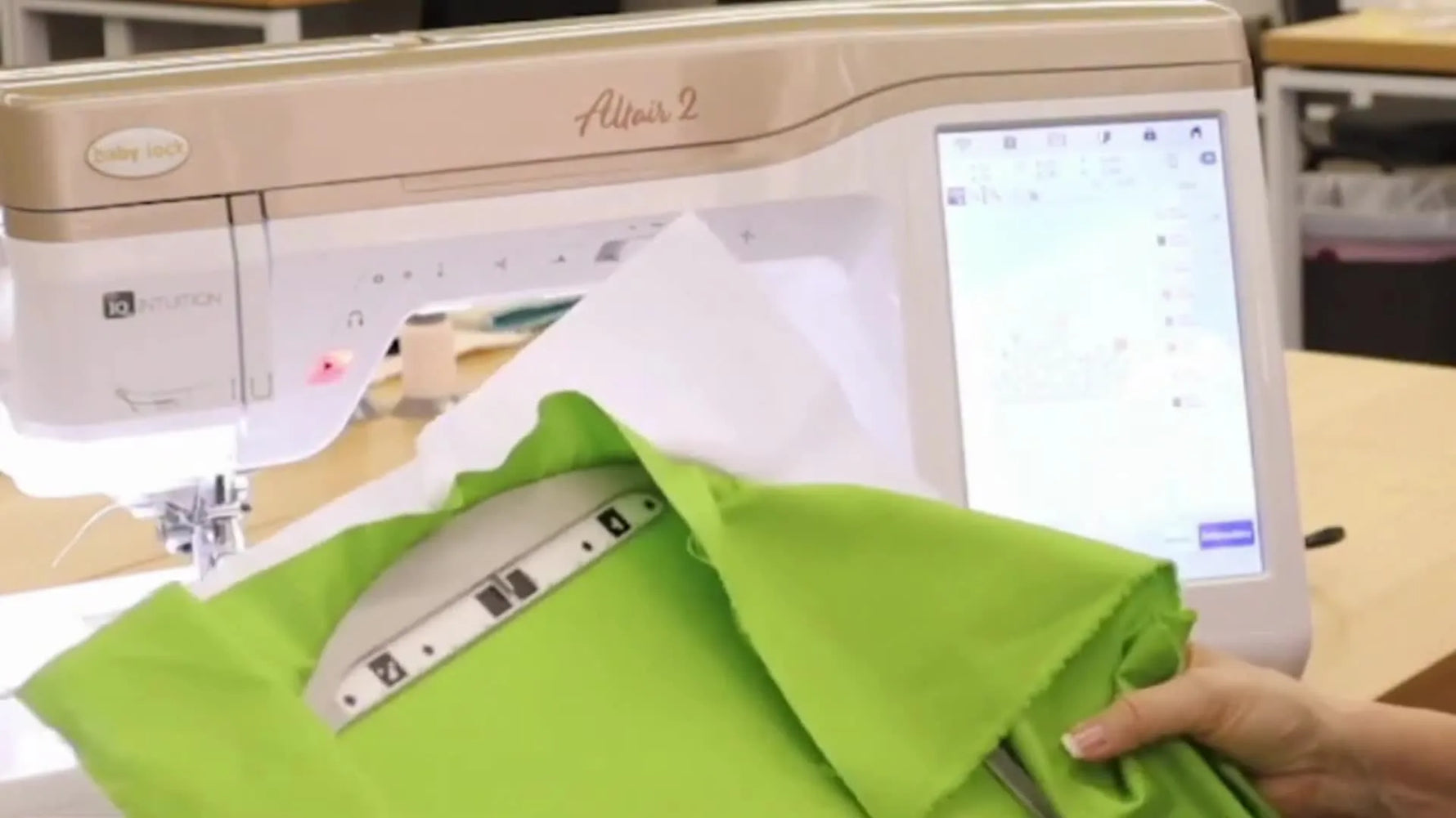 Denise holds a hooped green shirt and points to the positioning sticker used for Baby Lock Altair 2 2‑Point Positioning alignment.