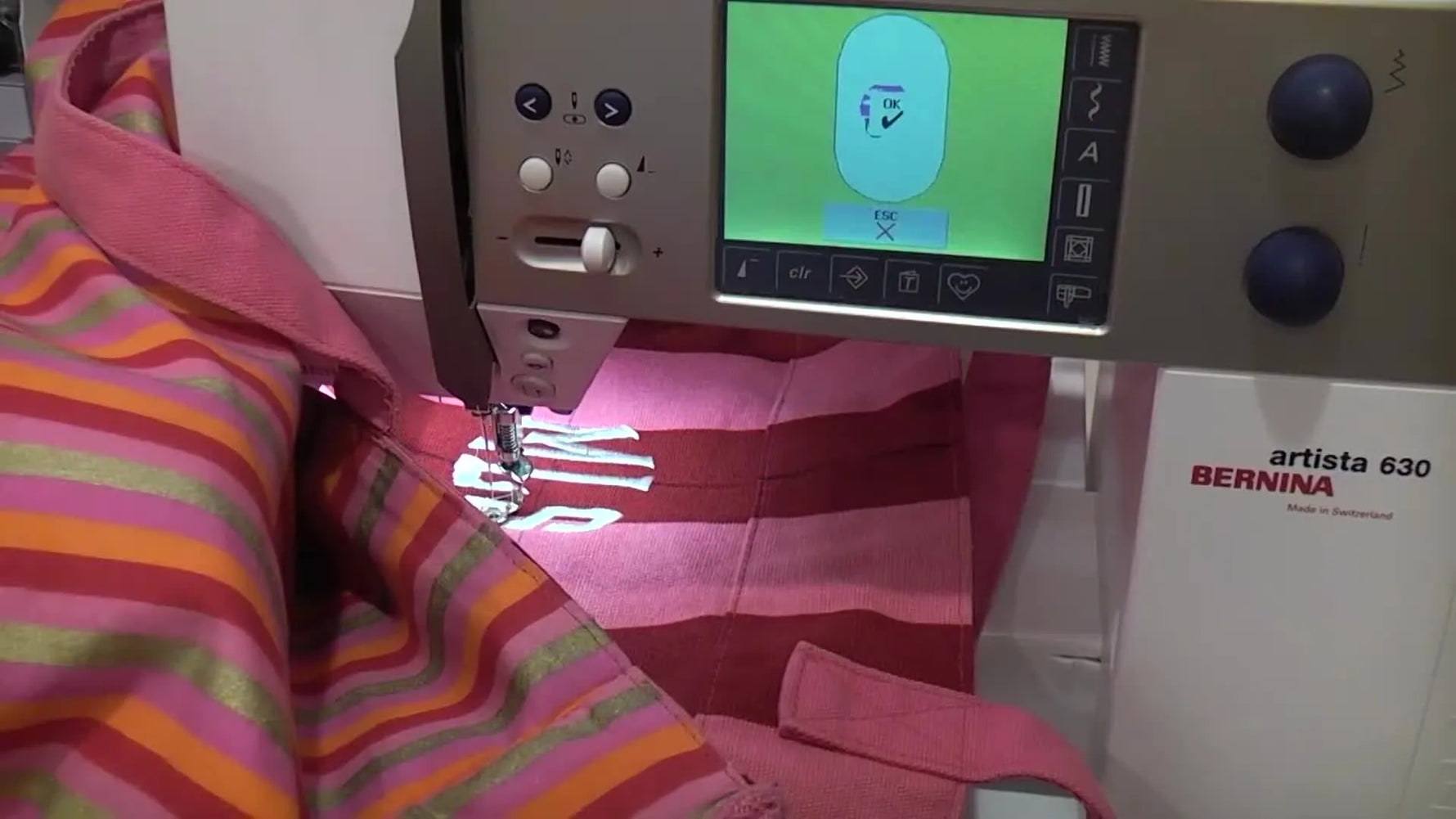 A close-up of the Bernina stitching a crisp white “MLS” monogram on a pink striped tote bag, showing the final placement and stitch quality.