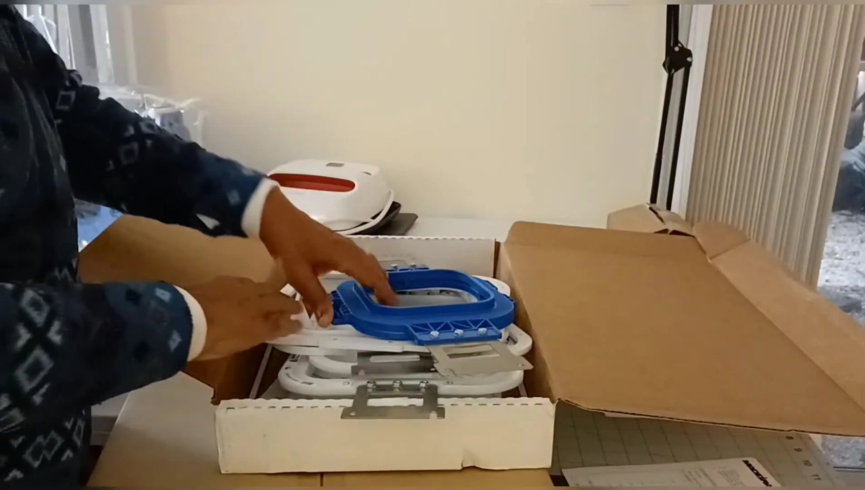 A large magnetic hoop with blue handles is lifted from the box, showing the brackets that mount it to a multi-needle embroidery machine.