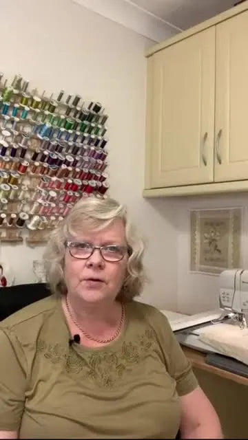 A wide landscape view of an embroidery studio with a large thread rack behind the presenter and an embroidery machine visible in the background.