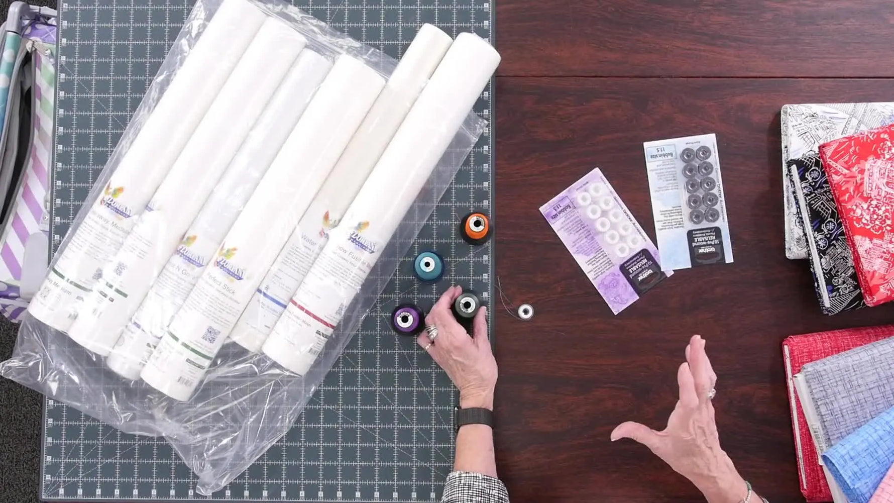 A wide tabletop view showing fabric bolts, thread spools, and bobbin packs laid out for choosing the right embroidery thread system.