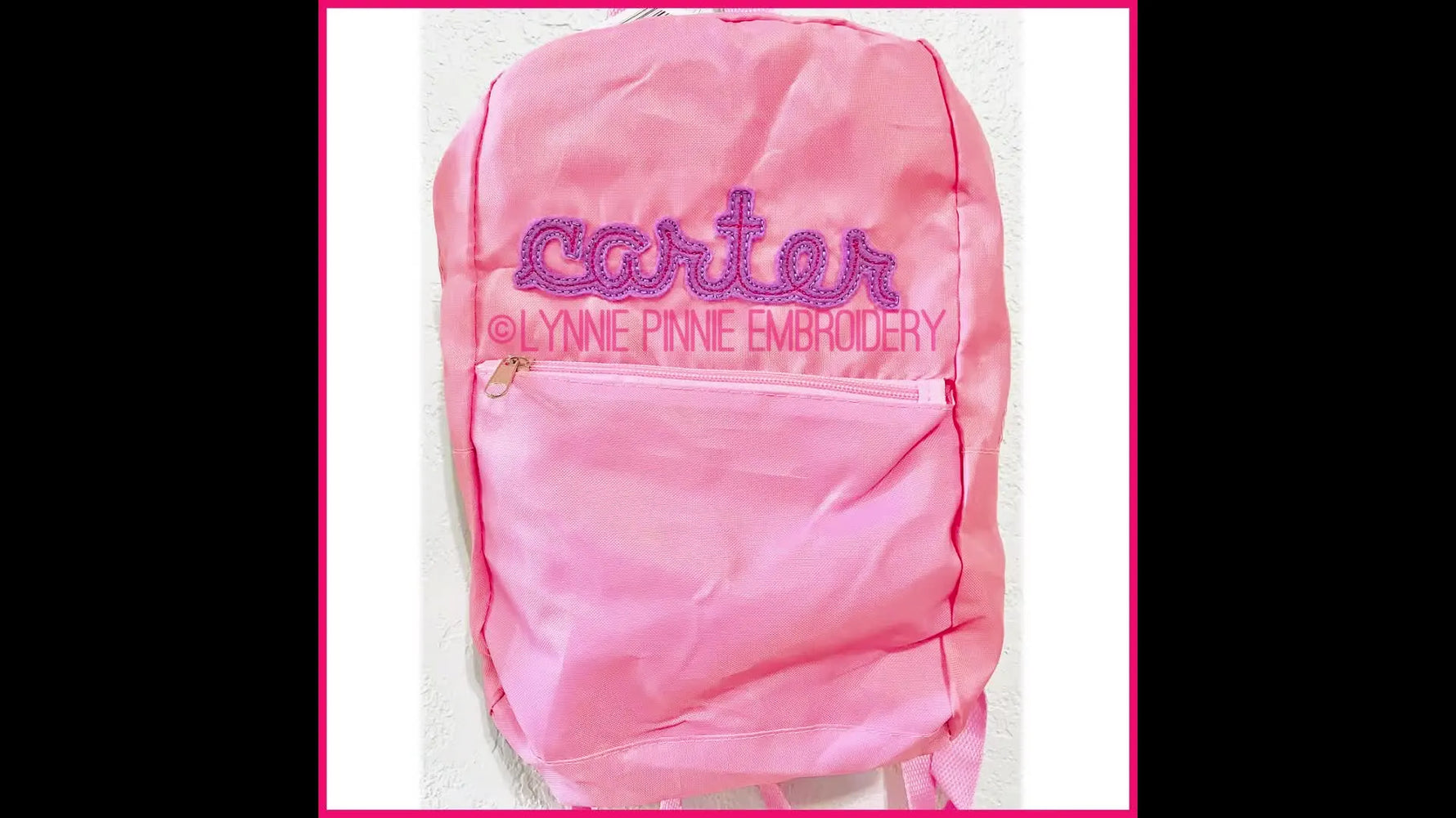A finished pink backpack hangs on a wall, showing a crisp raw-edge appliqué name that reads “carter.”