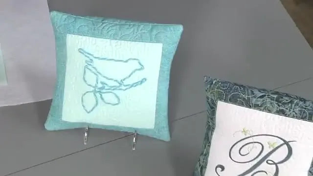 A finished teal in-the-hoop pillow cover stands upright, featuring a textured light-blue couched yarn bird on a quilted background.