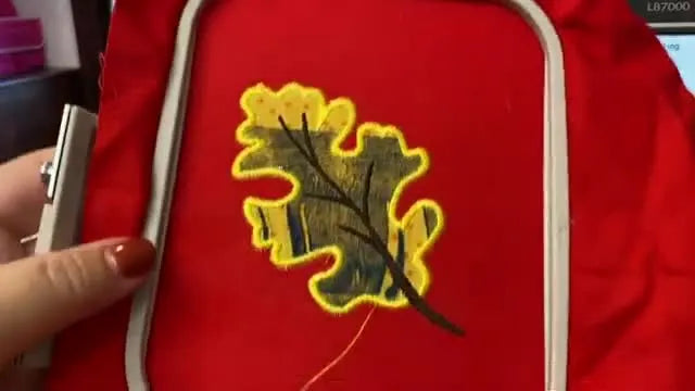 A finished leaf appliqué is held up in the hoop, showing a clean satin edge and a stem detail stitched on top.