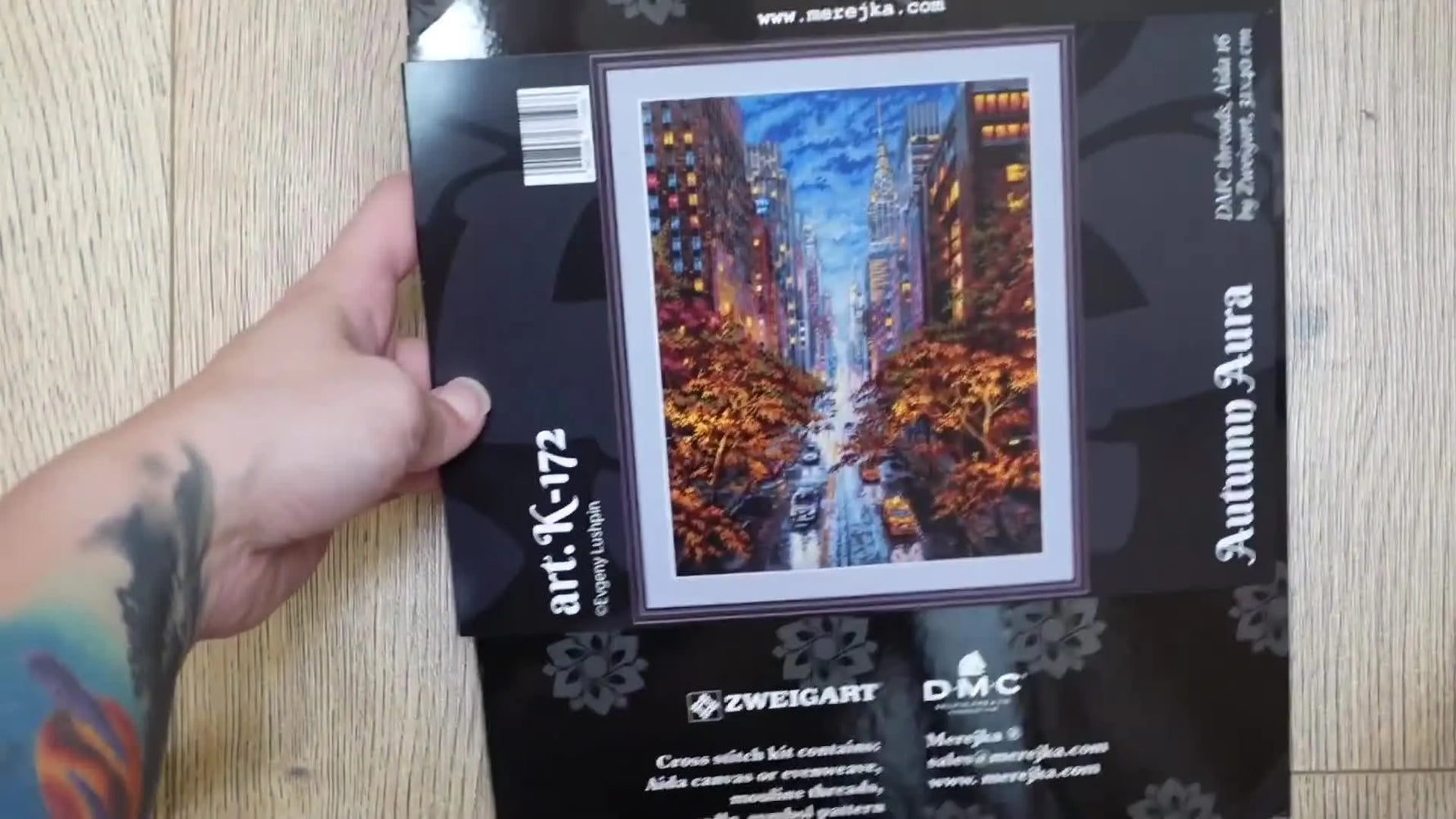 A woman's hands holding up a cross-stitch kit named 'Autumn Aura' by Merejka.