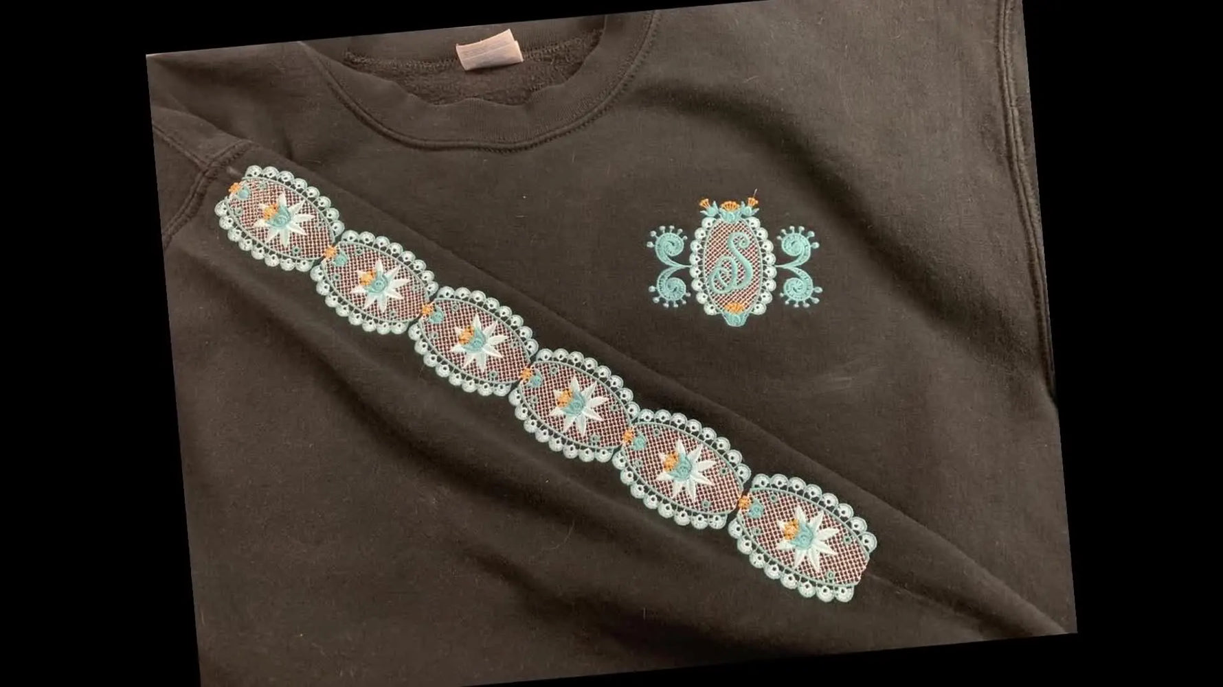 A close-up of a black sweatshirt sleeve stitched with continuous blue-and-white lace embroidery, showing crisp alignment and clean coverage.