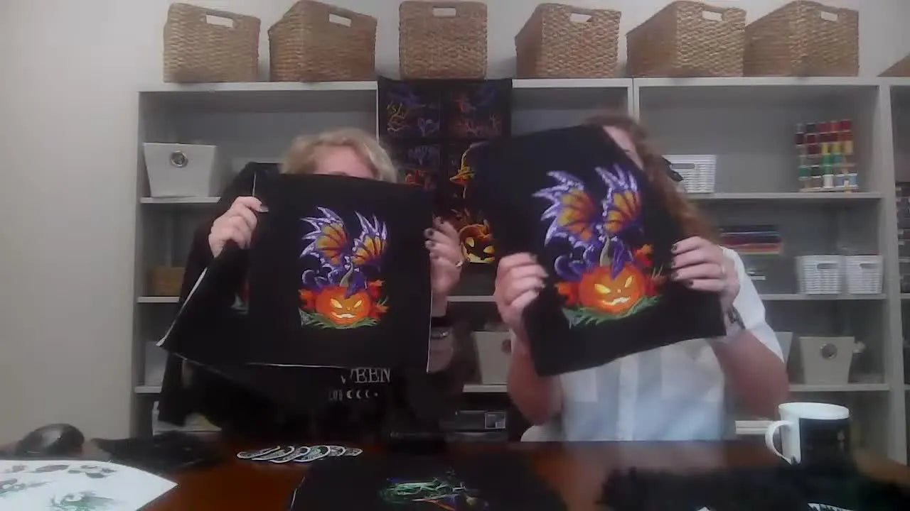 Two hosts hold up three different stitched sizes of the same Pumpkin Dragon design on black felt to show how sizing changes real stitch results.