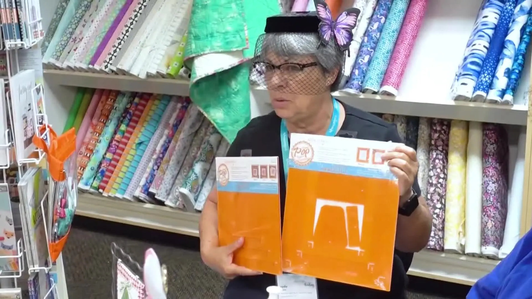 A host holds up Orange Pop Rulers packaging during a tool demo, highlighting the cutting-slot design used to square quilt blocks accurately.