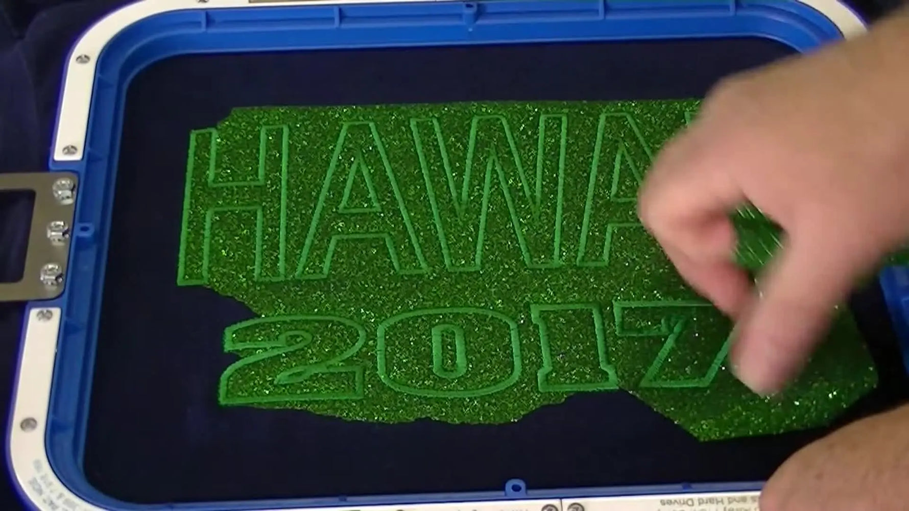 A close-up shows the excess green glitter sheet tearing away cleanly to reveal crisp satin-stitched “HAWAII” letters.