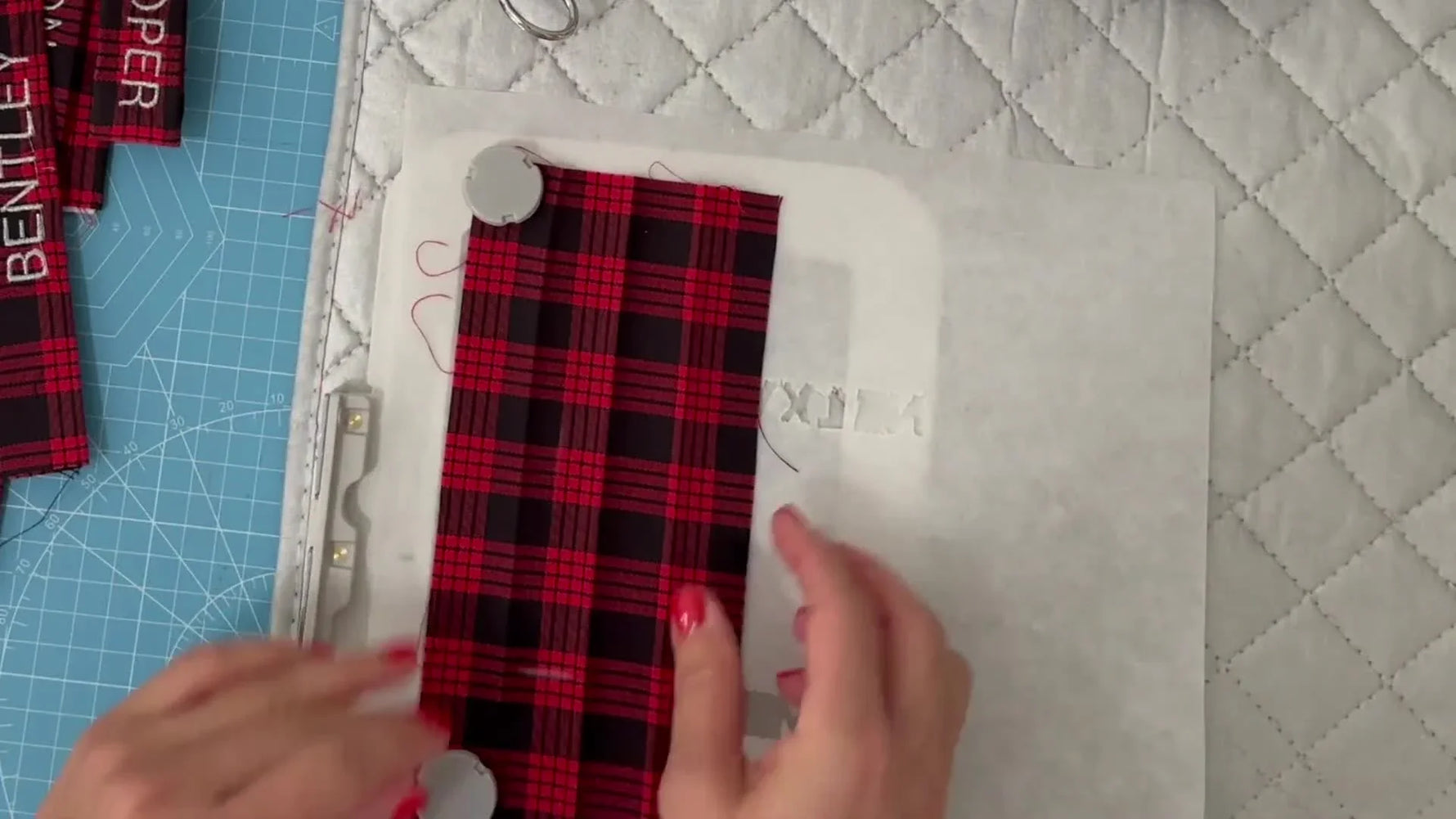 An overhead view of a 4x4 magnetic hoop holding a narrow plaid fabric strap flat on tearaway stabilizer while round magnets secure both ends for straight, clean embroidery.