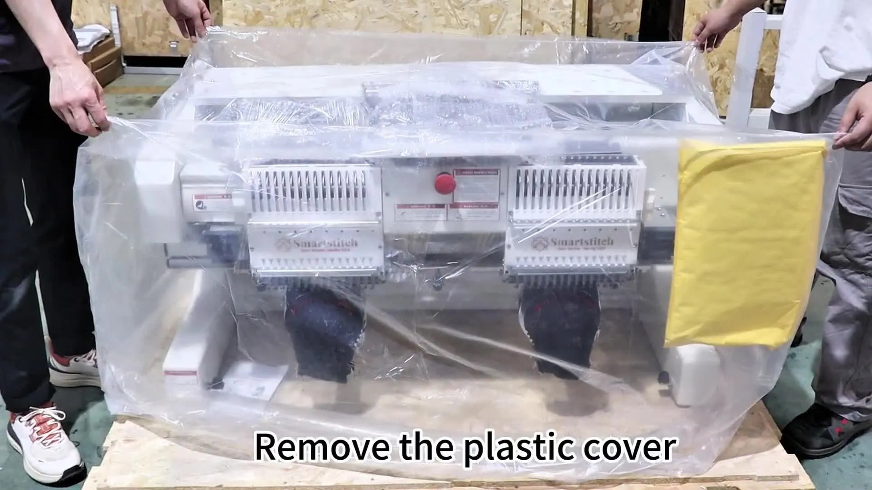 Two people lift the protective plastic cover off the Smartstitch S-1502HC, revealing the dual embroidery heads ready for installation.