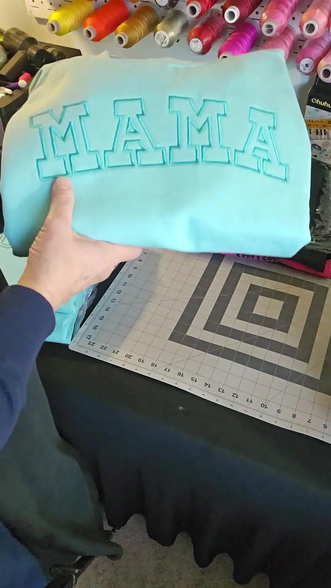 A teal Comfort Colors sweatshirt featuring a bold “MAMA” applique-style embroidery displayed as a high-impact gift and best-seller example.