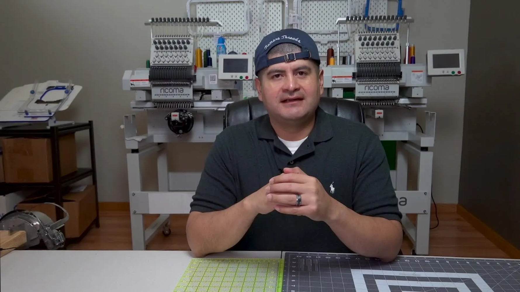 The instructor sits at a work desk with multi-needle embroidery machines behind him, framing the discussion around building a hat embroidery brand.