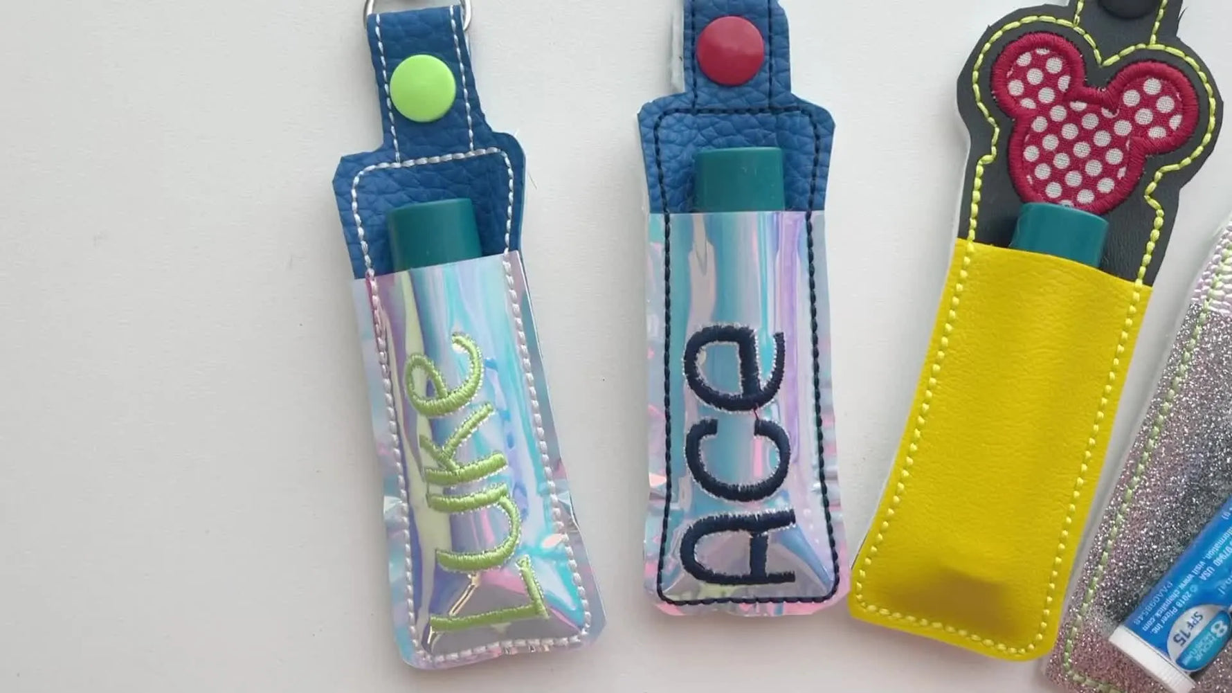 A flat lay of finished holographic and vinyl ITH lip balm holders with lobster clasps, showing multiple styles and personalization options.