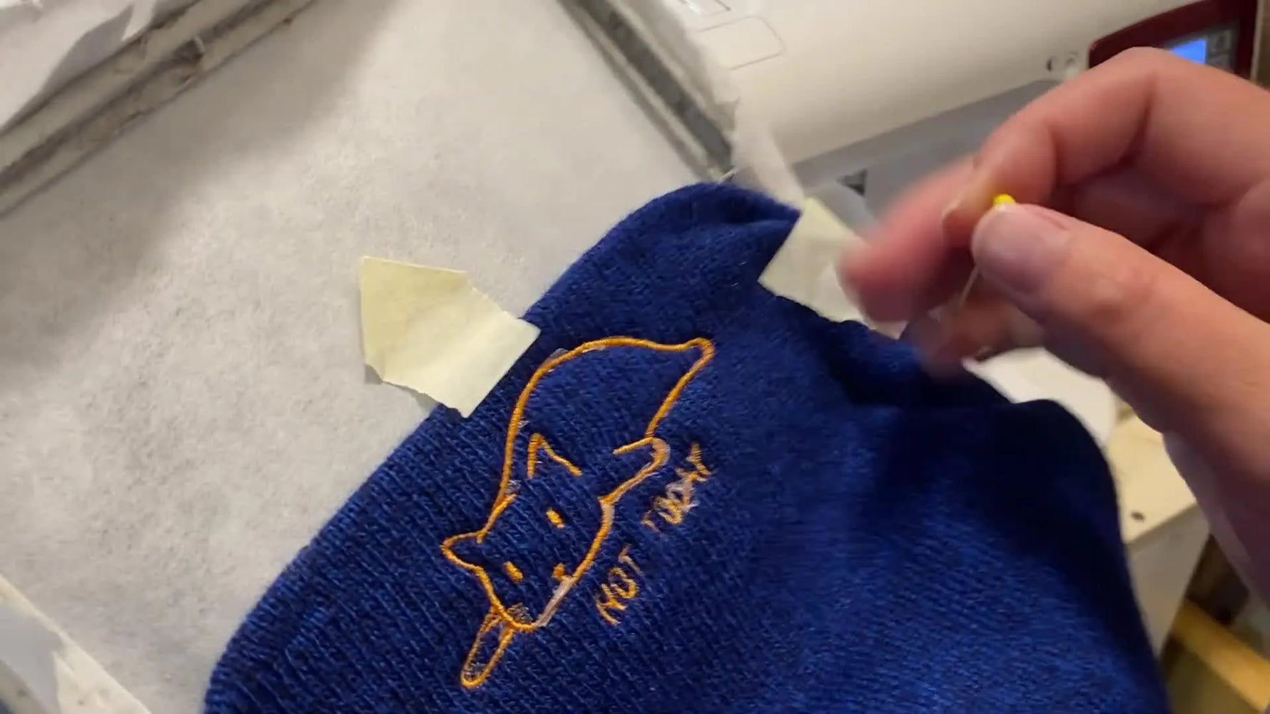 A finished blue knit beanie is held up to show a clean orange cat design with sharp lettering on the cuff.