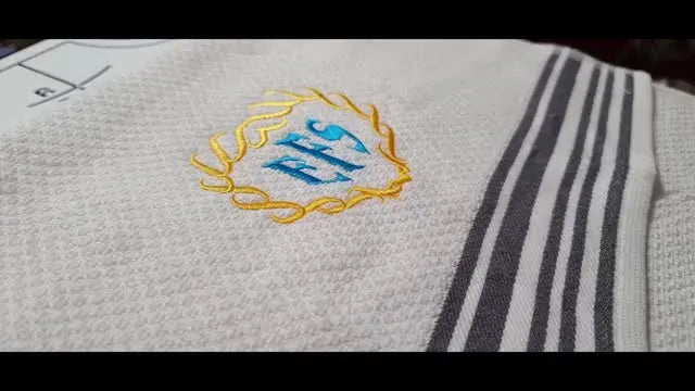 A close-up photo of the finished blue-and-gold three-letter monogram stitched on a white waffle weave towel.