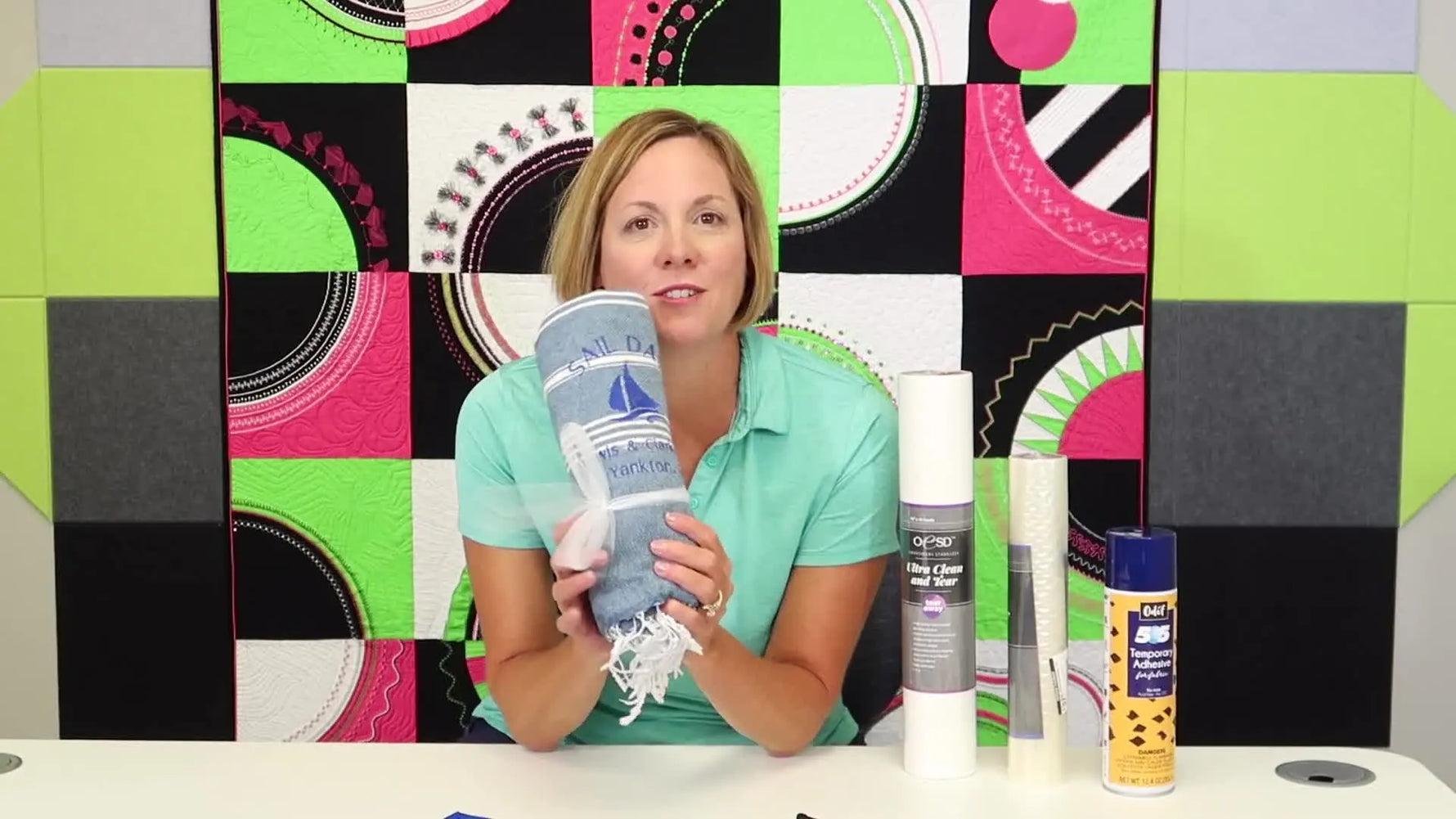 A finished Turkish towel with a crisp one-color sailboat logo sits beside stabilizer rolls and temporary adhesive spray, showing the full bulk-embroidery setup at a glance.